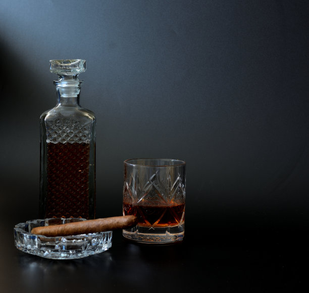 一个玻璃烟灰缸里的古巴雪茄，一个水晶酒瓶和一杯黑色背景的威士忌。图片下载