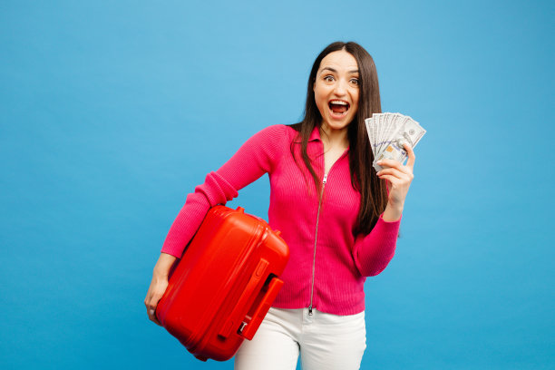 积极的女孩带着行李和钱要去旅行。穿着粉色毛衣和牛仔裤的女人站在蓝色背景上。假期的概念图片下载