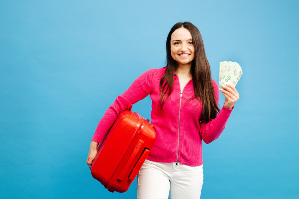 积极的女孩带着行李和钱要去旅行。穿着粉色毛衣和牛仔裤的女人站在蓝色背景上。假期的概念图片下载