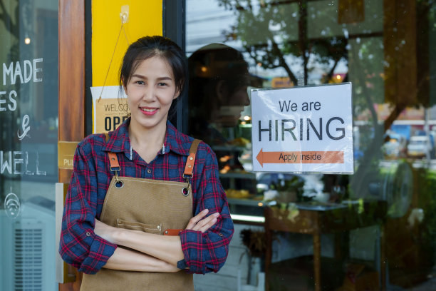 一位美丽的亚洲咖啡店老板的特写镜头。小户型的工作和招聘标志被贴在玻璃门上。咖啡店的女人站在店前，双臂交叉图片下载