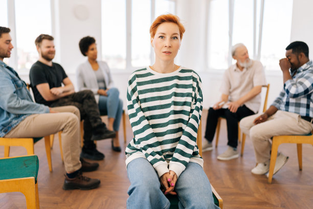 在集体治疗过程中，漂亮的年轻女子看着镜头的肖像，背景是多元文化和不同年龄的人围坐在一起。图片下载