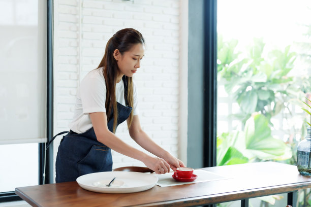 漂亮的亚洲咖啡馆女工。在家里的小咖啡店里收集盘子和玻璃杯，为下一批顾客清理桌子。这名妇女拥有一家小型家族企业。图片下载