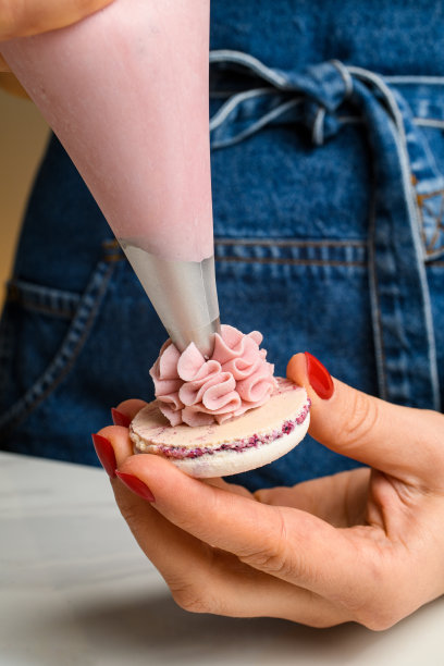 涂着鲜亮指甲的女厨师从糖果袋里挤出粉红色的水果奶油，放在马卡龙的外壳上。图片下载
