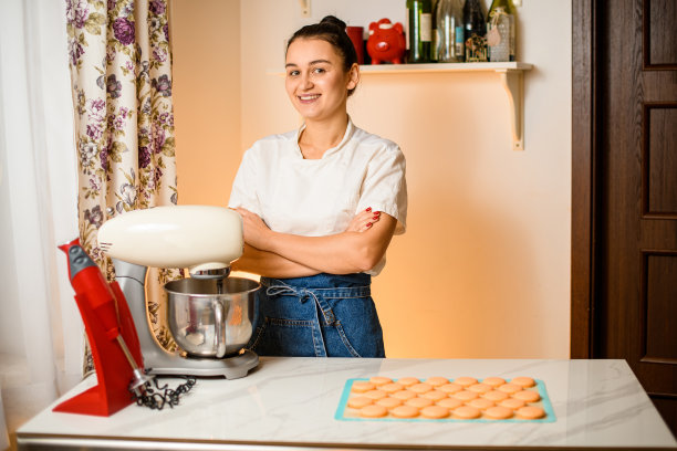 一个年轻的微笑着的女人站在厨房里拿着金属碗的食品加工机旁图片下载