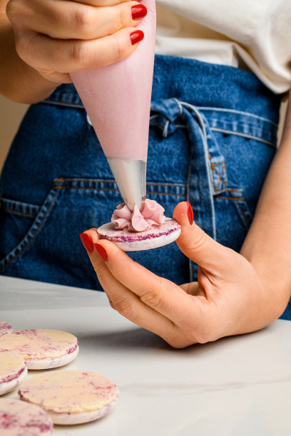 特写镜头，女厨师从马卡龙外壳上的糖果袋中挤出粉红色的水果甘纳许奶油。图片下载