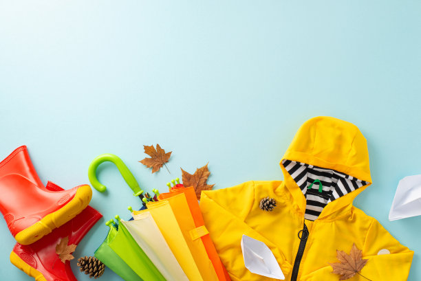 秋天的魔法。头顶布置展示儿童雨衣，胶鞋，彩虹设计的充满活力的雨伞，纸船，枫叶，松果，浅蓝色表面，提供文字或广告的空间图片下载