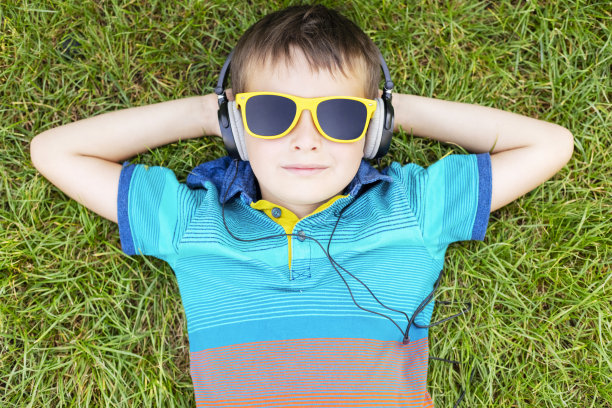阳光明媚的日子里，快乐的男孩戴着太阳镜躺在草地上，戴着耳机听音乐图片下载