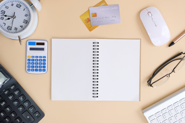 空白记事本和一支笔放在办公桌上，周围是电脑设备和各种纸张。平铺，俯视图，货币和银行背景图片下载