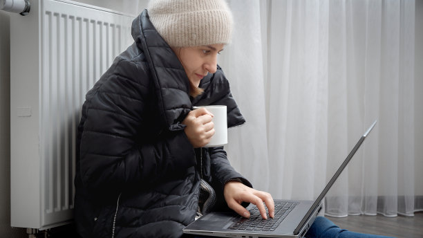 戴着帽子穿大衣的女人在家里靠暖气片用笔记本电脑工作。能源危机的概念，高账单，坏了的供暖系统，节约和节省每月的公用事业费用。图片下载