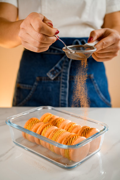 女糖果师用筛子将新鲜出炉的法式马卡龙撒上橙粉。前视图。图片下载