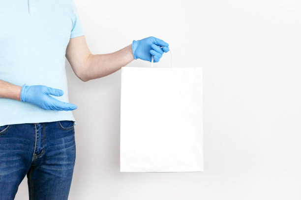 检疫期间交货的概念。戴蓝色手套的年轻人手里拿着白色的纸袋，背景是白色的。2019-nCov大流行中的非接触式递送图片下载