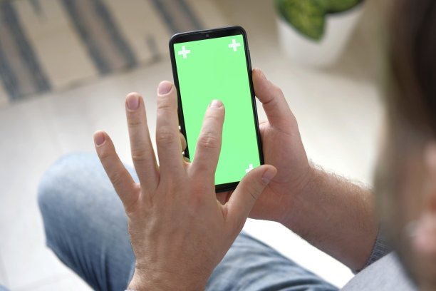 年轻人坐在家里，手里拿着智能手机的绿色模型屏幕。男性使用色度键手机。垂直模式。触摸，滑动显示屏，点击，浏览网络社交媒体图片下载