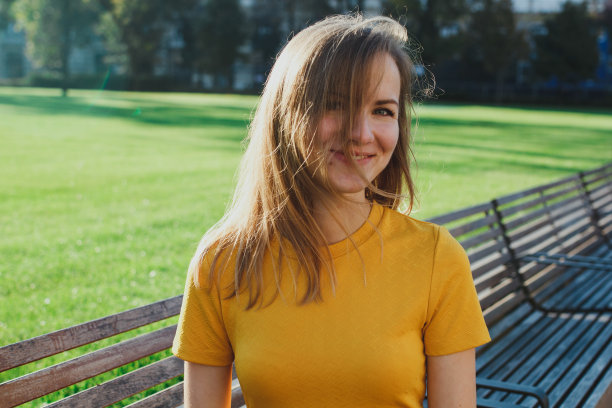 穿着芥末色连衣裙的漂亮女孩坐在绿油油的草坪旁的木凳上微笑着。阳光明媚的早晨照片。快乐的生活方式。背景是九月的树木。图片下载