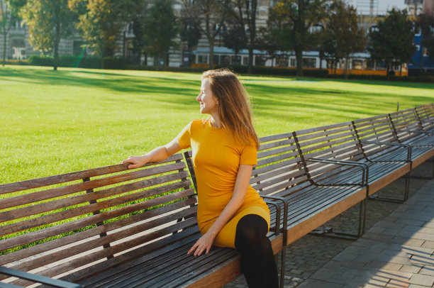 穿着芥末色连衣裙的漂亮女孩坐在绿油油的草坪旁的木凳上微笑着。阳光明媚的早晨照片。快乐的生活方式。背景是九月的树木。图片下载