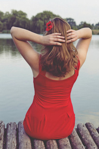 穿红衣服的漂亮女孩坐在木码头上。一个女人在湖边用红花挽着她的头发。乡村和自然的户外照片。夏天的河水，傍晚溅起的水花。图片下载