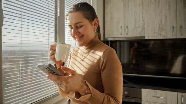 微笑的女人站在厨房的窗户边喝咖啡边使用智能手机。早晨的人们，美丽的城市风景，在办公室休息。图片下载
