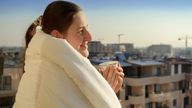 幸福微笑的女人端着一杯咖啡，从阳台上看着太阳从城市的天际线升起图片下载