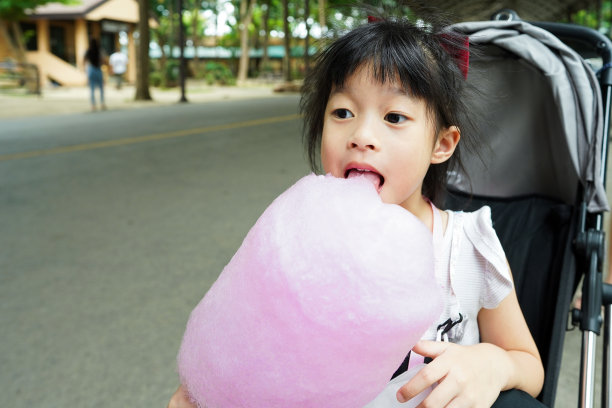 亚洲可爱的四岁小女孩坐在婴儿车里，拿着粉红色的棉花糖在户外吃棉花糖，太好吃了。图片下载