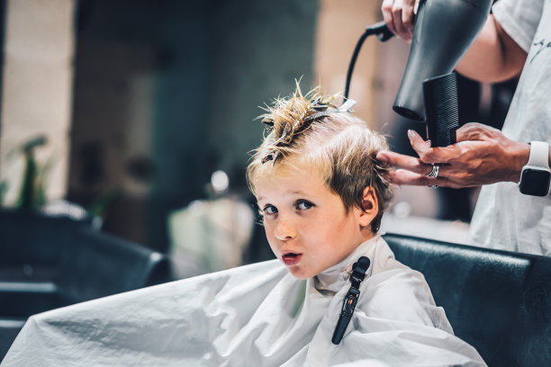 生活方式照片理发店的金发男孩顾客。美发师男士工作时动手梳子工具剪刀干衣机夹白色披肩。现代风格的儿童短发。为儿童提供时尚商业服务图片下载