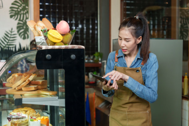 店主店是亚洲女人在店里点菜，站着和顾客聊天走进店里点面包房蛋糕，站着的女人微笑着和顾客打招呼，拿着托盘拿起托盘上的零食。图片下载