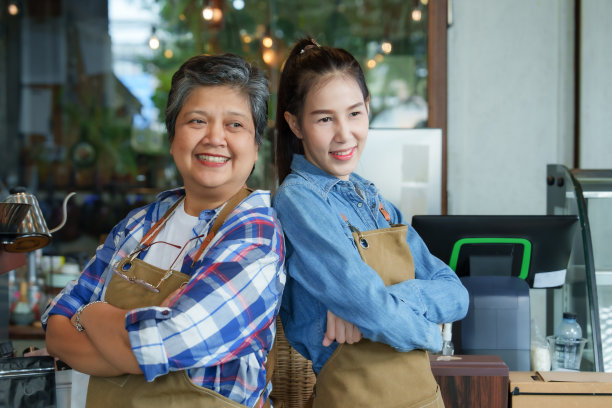 年迈的单身母亲抱着双臂站在美丽的女儿身边，眼中充满了幸福。每天早上准备开咖啡店，小家族生意。一个女人开了一家咖啡馆。图片下载