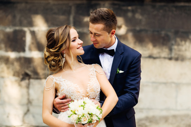 新郎望着以砖墙为背景捧着结婚花束的新娘图片下载