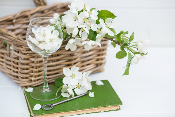 美丽静物，春光卡。一棵苹果树上的白花躺在一个玻璃杯里，周围环绕着鲜花，一个柳条筐和一张旧的绿纸。水平照片与复制空间。白色背景。图片下载