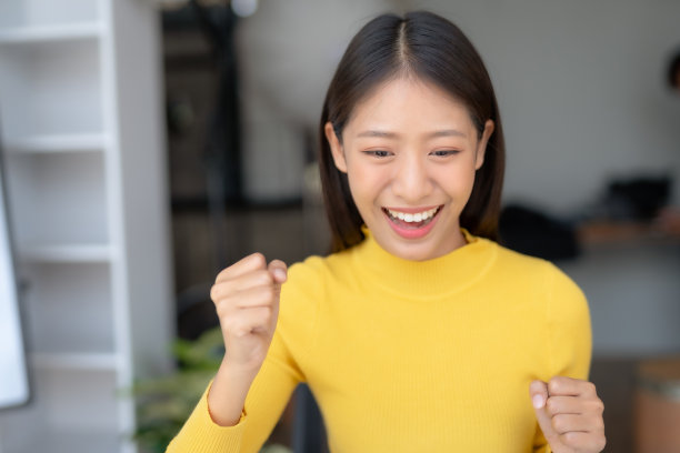 成功的亚洲女商人喜欢家里有一台笔记本电脑。自由职业者亚洲女人大喊“是”，看着笔记本电脑屏幕，庆祝胜利，目标实现举起手臂。图片下载
