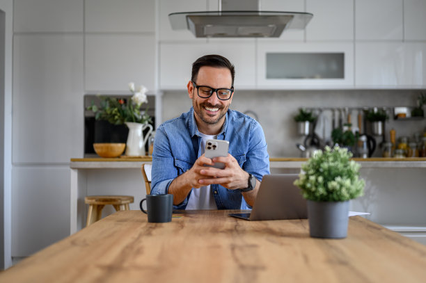 微笑的年轻男性企业家在家庭办公室的桌子上用笔记本电脑用手机聊天图片下载