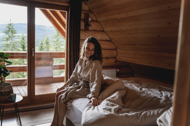 一位年轻的白人女子坐在山间小木屋的床上，抚摸着床单。舒适的睡眠在一个木屋概念图片下载