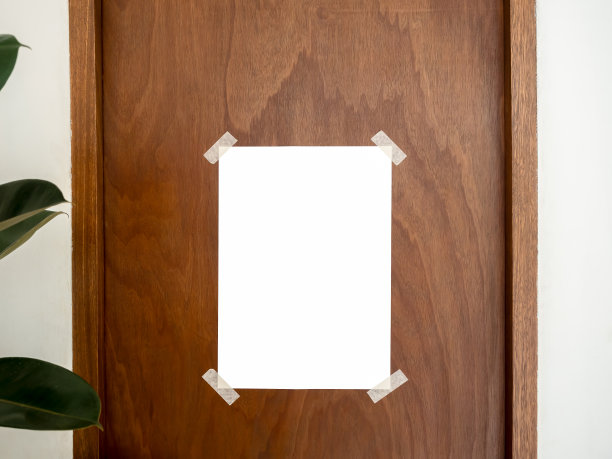 原型空白色垂直海报贴在复古棕色木门和白色墙壁背景附近的绿叶在咖啡馆，最小的风格。空白海报模板与胶带在角落，为广告。图片下载