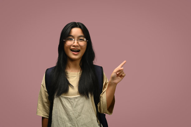 微笑的亚洲青少年学生用手指指着粉红色背景上羞涩的空间隔离图片下载