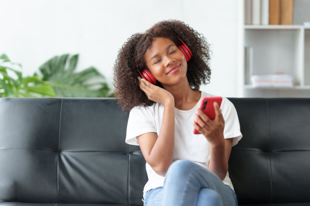 明亮可爱的亚洲年轻女子戴着耳机坐在沙发上，用智能手机听音乐，在家听音乐，快乐放松。图片下载