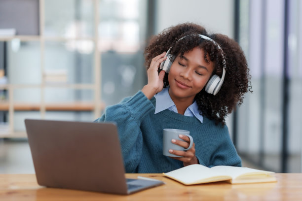 快乐的年轻女子，一头卷发，戴着白色耳机，拿着咖啡杯，在笔记本电脑上听音乐。图片下载