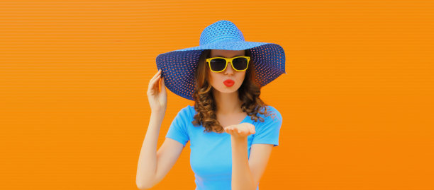 夏天的肖像，美丽的年轻女子吹着她的嘴唇送甜蜜的飞吻，戴着橙色背景的草帽图片下载