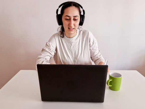 漂亮的年轻女人在家里用着笔记本电脑，戴着耳机，喝着饮料图片下载