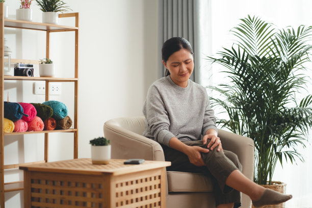 患有膝盖疼痛的亚洲妇女坐在家里客厅的扶手椅上。图片下载