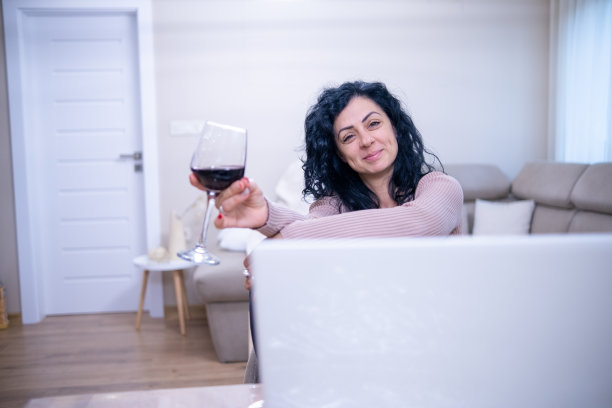一位年轻女子坐在笔记本电脑前，手里拿着一杯红酒。图片下载