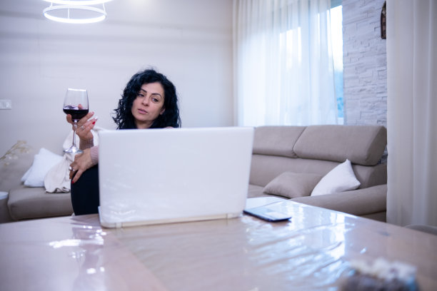 一位年轻女子坐在笔记本电脑前，手里拿着一杯红酒。图片下载