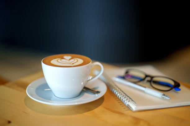 一张木桌上一杯咖啡的特写图片，背景是记事本、笔和眼镜，复制空间。图片下载