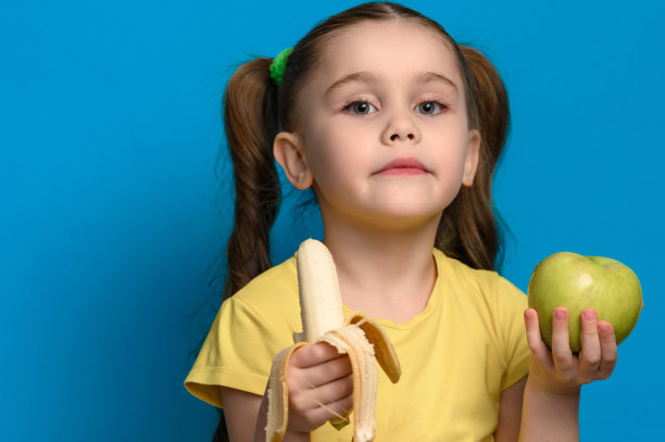 一个孩子手里拿着一个青苹果和一根香蕉，背景是蓝色的，一个女孩代表健康和健康的食物。图片下载