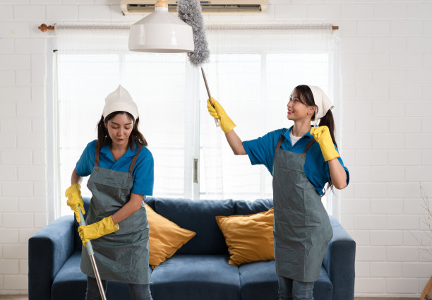 快乐的两个亚洲妇女穿着工作服女仆打扫家里，手里拿着除尘刷，用拖把在家里的厨房里擦拭图片下载