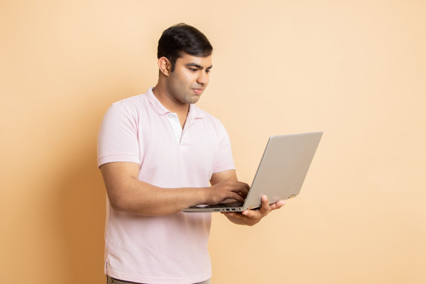 穿着便服的年轻印度成年男子手里拿着笔记本电脑，孤立在米色的工作室背景下。人们的生活方式，盖伊使用电脑。图片下载