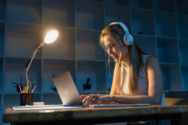 年轻快乐的亚洲妇女戴着白色耳机听音乐，晚上在笔记本电脑上做她的项目图片下载