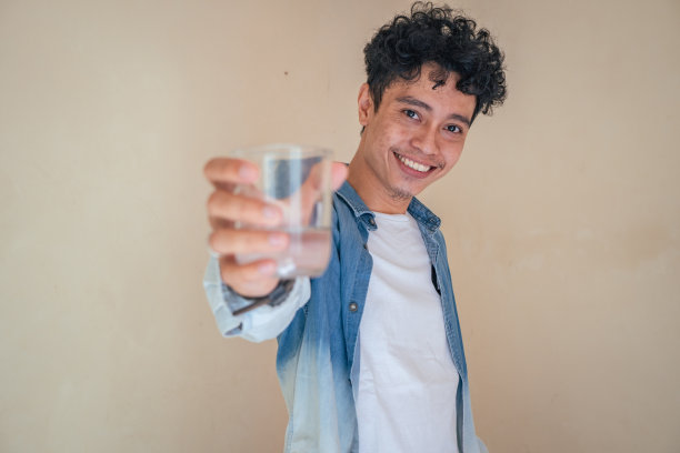 穿着牛仔衣服的年轻卷发男子端着一杯水。微笑，欢呼和快乐的表情。图片下载