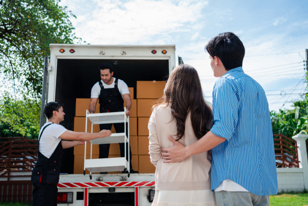 一对亚洲夫妇一边检查，一边把箱子和家具从一辆小货车上卸到一所有服务货物的新房子里，两名穿着制服的搬运工搬运箱子。搬家和送货的概念。图片下载