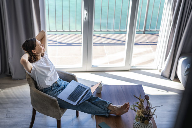 放松快乐的年轻女子坐在椅子上，腿放在咖啡桌上，在家里使用笔记本电脑冲浪，在线工作，电子商务和购物概念图片下载