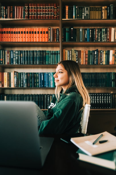 年轻女大学生的肖像坐在书架附近，用笔记本电脑学习，准备考试，写论文做家庭作业在家里。图片下载