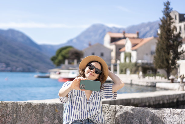 在地中海度假胜地的海滩上，一位戴着帽子微笑着用智能手机自拍的年轻女子图片下载