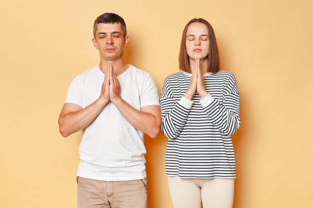 一个穿着休闲服的放松的男人和女人站在米色的背景上，闭着眼睛做着祈祷的姿势，一起练习瑜伽。图片下载